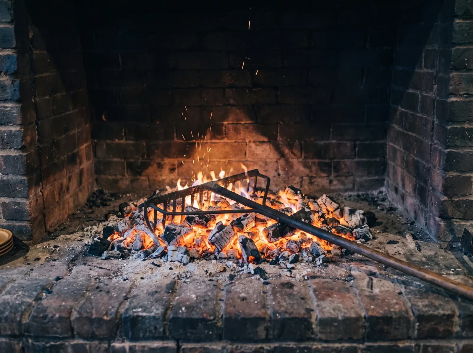A long iron coal rake resting in glowing birch coals inside the brick hearth, sparks lifting