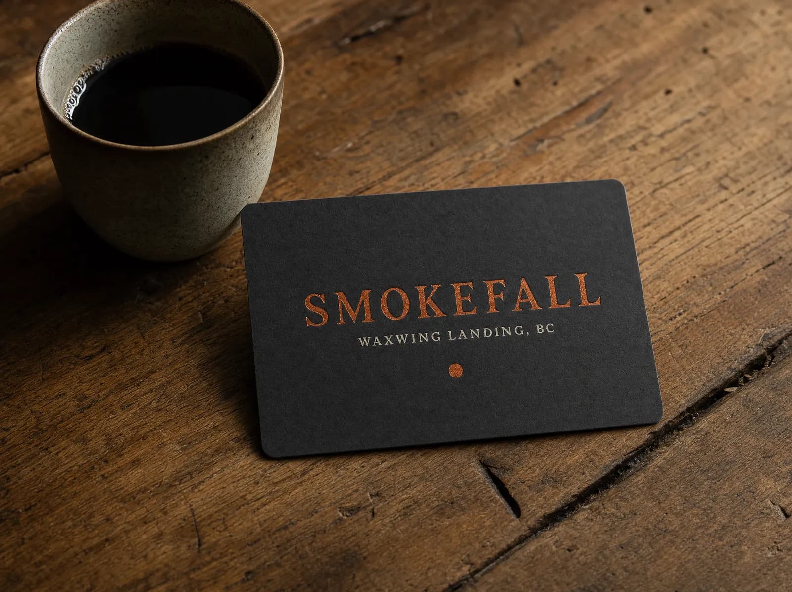 The Smokefall gift card on dark board stock, ember-orange lettering, lying on a wooden table beside a coffee